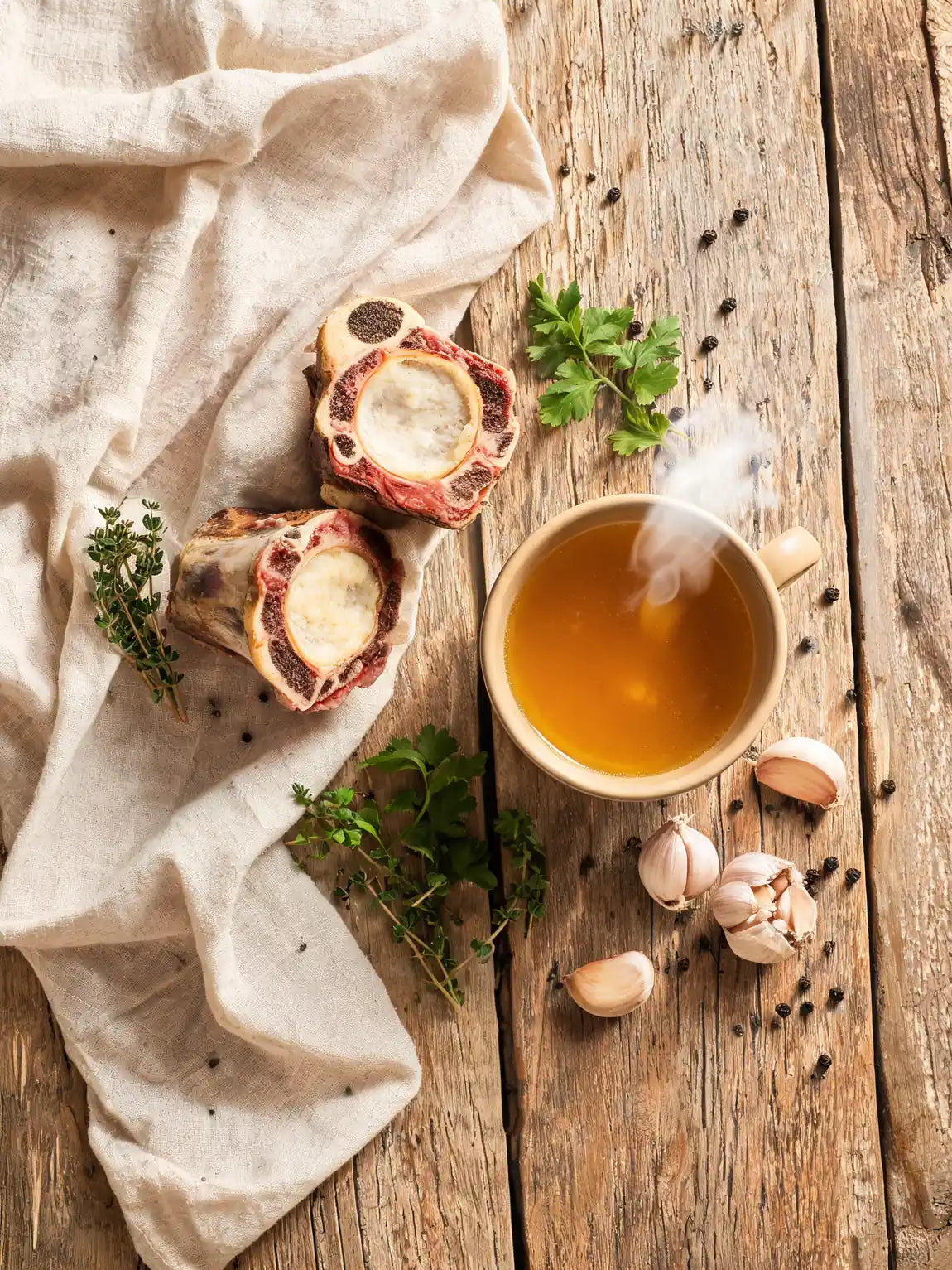 Grass-fed beef marrow bones, fresh herbs and a steaming mug of golden bone broth on a wooden board
